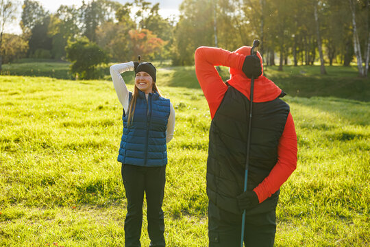 A smiling woman and a man in outdoor gear stretch in a sunny grassy field, preparing for a Nordic walking session with poles, surrounded by trees and open nature