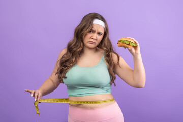A sad young european plus size woman dressed in sportswear holds a burger in one hand while measuring her waist with a tape measure in the other.