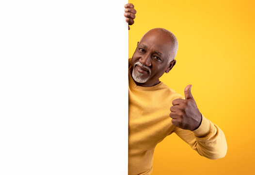 A cheerful senior black man stands in front of an orange background, holding a blank white paper banner with one hand. He gives a thumbs up gesture, inviting ad designs for display.