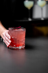 A close-up of a person's hand holding a pink cocktail in a patterned glass on a dark, glossy bar counter. The drink is garnished with a flower petal.