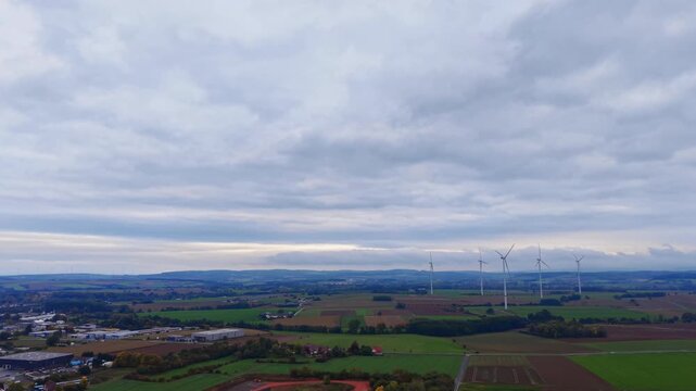 Vast green fields blend with patches of farmland, dotted with wind turbines standing tall against a cloudy sky. Tranquility echoes in this beautiful rural landscape.