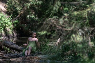 Young woman with short hair, dressed in a green wrap, sits gracefully by a serene riverbank surrounded by lush greenery, embodying tranquility and connection with nature