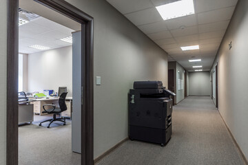 A clean, modern office hallway features a printer beside an open door leading to a workspace. Cubicles are visible, creating a productive atmosphere. The space is well-lit and organized.