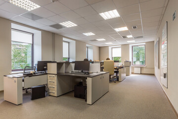 A well-lit office space shows several employees engaged in their tasks at desks with computers. Large windows provide natural light and a view of greenery outside.
