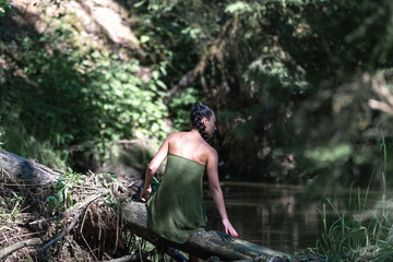 Young woman with short hair, dressed in a green wrap, sits gracefully by a serene riverbank surrounded by lush greenery, embodying tranquility and connection with nature