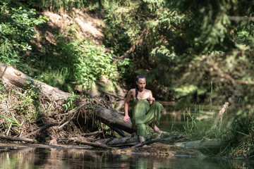 Young woman with short hair, dressed in a green wrap, sits gracefully by a serene riverbank surrounded by lush greenery, embodying tranquility and connection with nature
