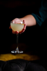 A hand in a dark green sleeve holds a coupe glass filled with a light-colored cocktail, garnished with a citrus twist, against a dark, moody background. The focus is on the glass.