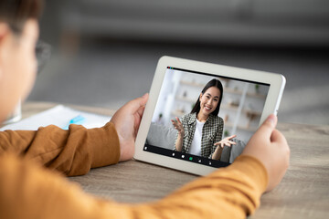 A cheerful young teacher guides an unrecognizable child during an online class using a digital tablet. The setting is cozy, creating a friendly learning environment at home.
