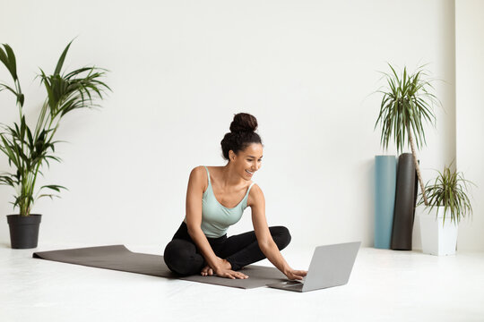 Sporty young woman enjoys selecting yoga video tutorials on her laptop. She is sitting on a fitness mat in a well-lit room surrounded by plants, ready for her workout.