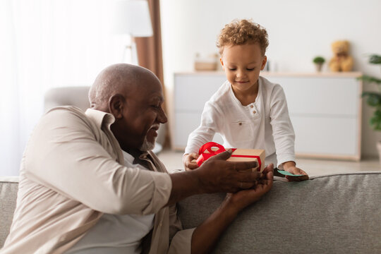 An African American grandfather joyfully presents a wrapped gift to his young grandson on his birthday. They are sitting together on a sofa in a cozy home environment, celebrating family joy. - Powered by Adobe