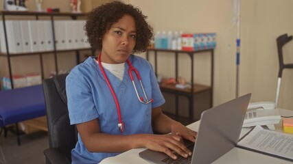 Woman doctor wearing blue scrubs and red stethoscope typing on laptop at desk in clinic; compassion dedication expertise focus. - Powered by Adobe