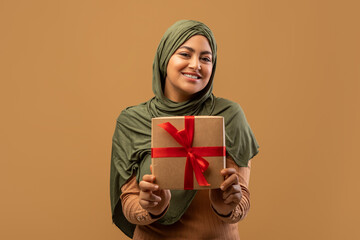 Cheerful arab lady in hijab holding gift box and smiling to camera, positive muslim woman with woman's day or birthday present posing over beige studio background