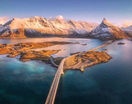 Aerial view of bridges, Reine village surrounded by fjords and snowy mountains at sunrise. Lofoten, Norway. Bright winter sunlight over fishing cabins, blue sea, and coastal landscape. Top drone view - Powered by Adobe