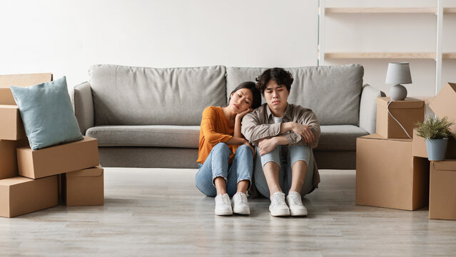 Exhausted young Asian couple sits on the floor among carton boxes, resting from the tiring task of unpacking on their moving day. The empty room creates a sense of fresh beginnings.