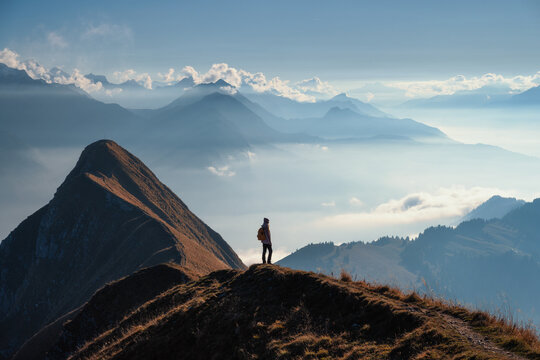 Woman with backpack standing on a mountain ridge above clouds in the Swiss Alps at sunset. Scenic view of alpine peaks, girl, mist, clear sky in fall. Concept of hiking, travel, and outdoor adventure