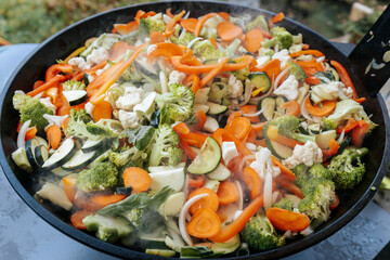 A vibrant mix of steaming stir-fried vegetables including carrots, broccoli, zucchini, cauliflower, and bell peppers, cooking in a large black pan outdoors.