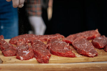 A person wearing gloves and a plaid shirt seasons raw steaks with ground pepper on a wooden board during an outdoor cooking demonstration.