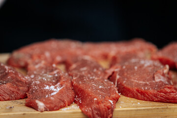 A person wearing gloves and a plaid shirt seasons raw steaks with ground pepper on a wooden board during an outdoor cooking demonstration.