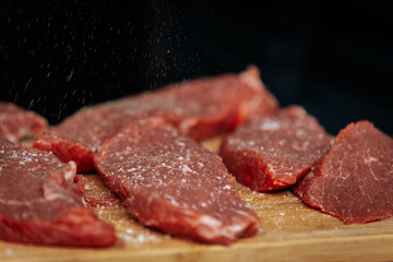 A person wearing gloves and a plaid shirt seasons raw steaks with ground pepper on a wooden board during an outdoor cooking demonstration.