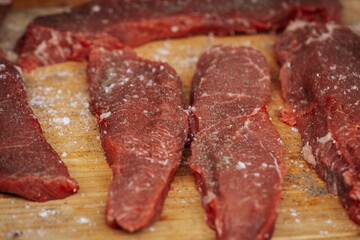 Close-up of raw beef steaks seasoned with salt and pepper, laid out on a wooden cutting board, ready for grilling at an outdoor cooking event.