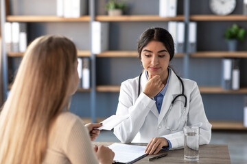Fototapeta premium A serious millennial Indian woman therapist in uniform reflects on an envelope containing money from a client in a clinic office. The setting is professional, highlighting healthcare ethics.