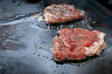 Four juicy steaks sear on a sizzling hot flat grill beside open flames, cooking in their own juices with a perfect crust forming on the surface.