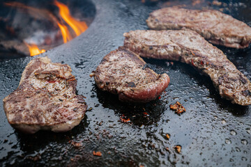 Raw steaks sizzle in a circle on a hot flat grill surrounding an open wood fire, capturing a dramatic top-down view of live-fire cooking in progress.
