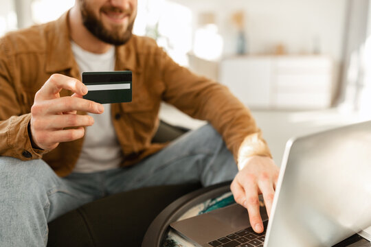 A smiling man sits on a couch, using his laptop to make an online purchase. He holds a credit card ready, enjoying a relaxed atmosphere at home.