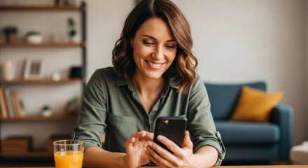 A smiling woman in her 30s wearing a green shirt, sitting at a wooden table with a glass of orange juice and using her smartphone, looking happy and relaxed