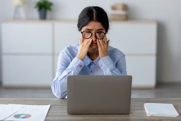 A tired young Indian business woman is seated at her desk, rubbing her eyes in discomfort. She appears stressed and fatigued as she struggles with her laptop amid a busy work environment.
