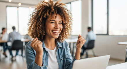 Young woman with curly hair is celebrating success while working on a laptop in a modern office environment, showcasing joy and achievement