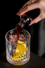 A hand pours a red-colored cocktail from a bottle into a textured glass filled with ice and a fresh orange slice, creating a dynamic, beverage-focused scene