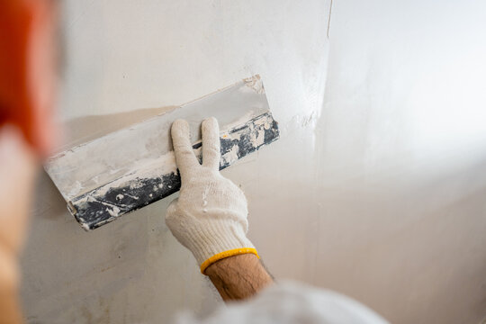 Close-up of a worker's gloved hand actively spreading and smoothing fresh plaster or joint compound onto an interior wall surface.
