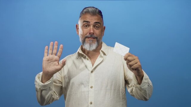 Bearded middle age man holding creditcard and raising palm in blue studio setting; skepticism caution.