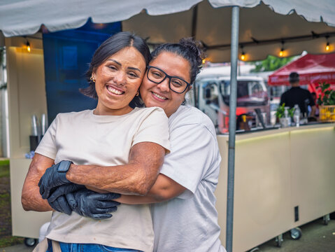 Portrait of a lesbian couple of entrepreneurial women in front of their street michelada stand, smiling and tending to their business.