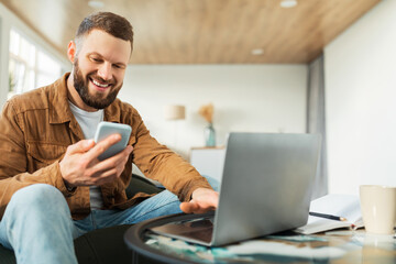 A young man sits comfortably on a couch, smiling at his phone while using a laptop on a coffee table. The relaxing atmosphere features modern decor and natural light.