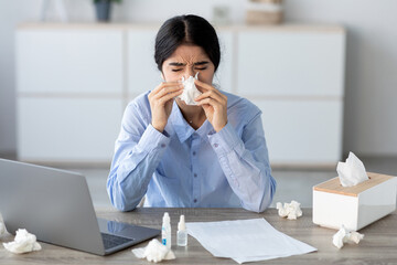 A young Hindu female manager is sitting at her home office desk. She blows her nose in a napkin,...