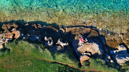 A lone figure in red stands on rugged coastal rocks, overlooking a vibrant turquoise sea. The scene...