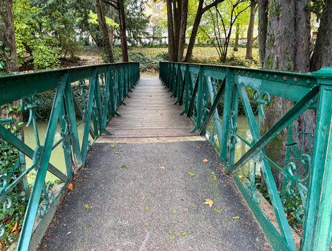 Old-fashioned wooden and iron pedestrian bridge with decorative railings over river in park, autumn scenery small town landscape, peaceful relaxing calm natural atmosphere with autumn leaves on path