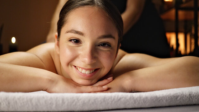 A smiling woman lies on a massage table, hands clasped under her chin. The serene spa setting emphasizes relaxation, wellness, and self-care, with soft lighting and comfortable posture.