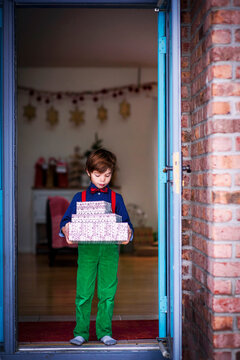 Young Boy in smart clothing standing in an open doorway holding a stack of wrapped Christmas gifts