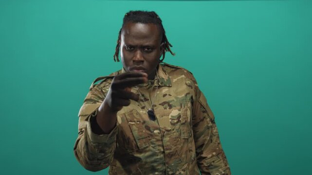 Man soldier in camouflage uniform pointing with hand and showing dog tags in teal studio; duty determination.