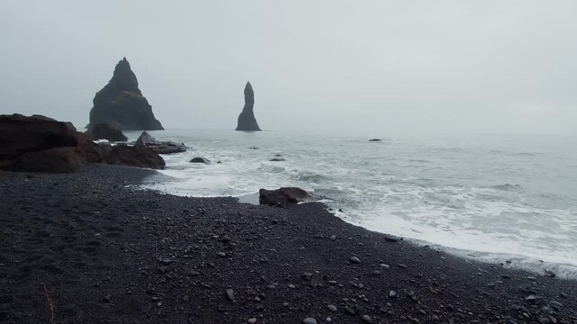 A moody and cinematic shot of the famous Reynisfjara black sand beach on the South Coast of Iceland. The powerful waves of the Atlantic Ocean crash against the unique black volcanic sand, with the ico