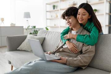 A young Asian couple relaxes together at home. The man is focused on his laptop while his wife hugs him, creating a cozy atmosphere.