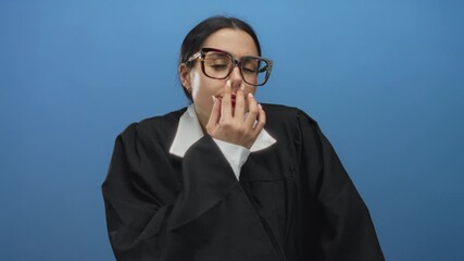 Woman wearing judge uniform yawning with hand over mouth against blue wall, conveying surprise and fatigue, displaying a thoughtful expression.