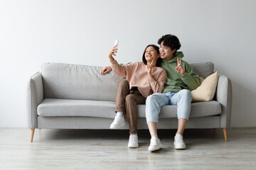 Obraz premium Joyful young man and woman sit on a gray sofa in their living room, smiling and taking a selfie with a smartphone. The atmosphere is warm and relaxed, showcasing their happiness together.