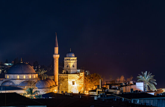 The historic Antalya Clock Tower and Tekeli Mehmet Pasha Mosque illuminated against the dark Mediterranean night, creating a stunning contrast between ancient architecture and sea. Antalya, Turkey.


