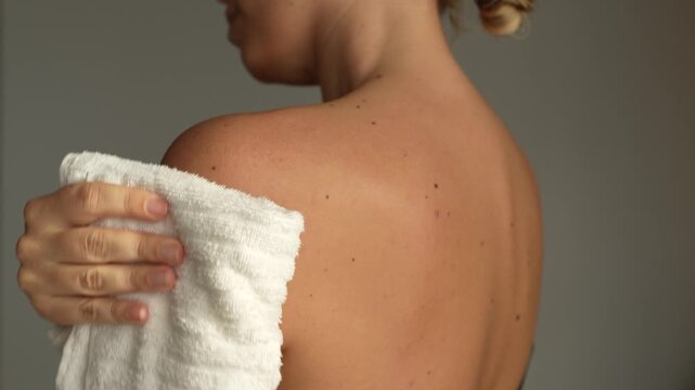 Young woman wiping sweat from shoulder and back after training on a light grey background. Concept of body cooling, sport recovery or hyperhidrosis awareness in natural light