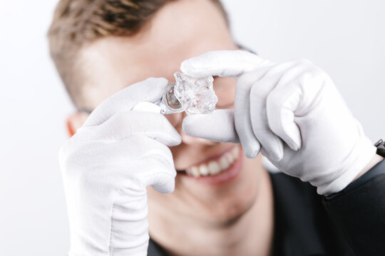 Happy Caucasian male jeweler examining large diamond with loupe and white gloves, white studio background - Powered by Adobe