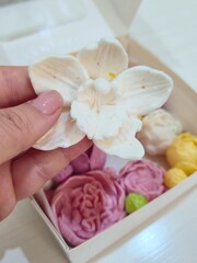 ​Close-up of a hand holding a white decorative orchid-shaped marshmallow or zephyr above a box of colorful floral treats